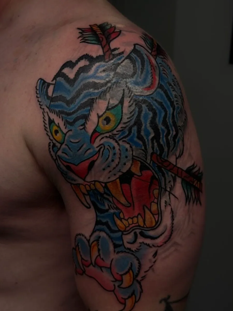 The same upper arm after the cover-up: a roaring blue Japanese tiger pierced by an arrow, tattooed over the old piece by Reed Drury at True North Tattoo in Lancaster, PA